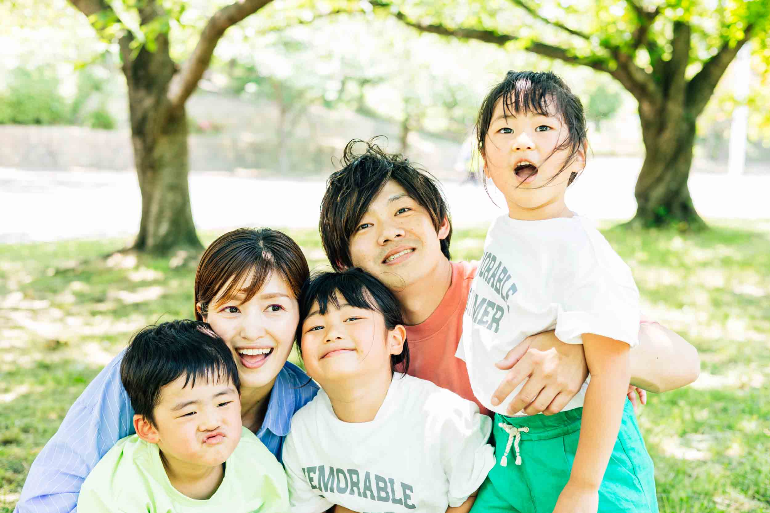 Family of five posing for a cheerful outdoor photo in a sunny park under green trees; everyone smiles at the camera.