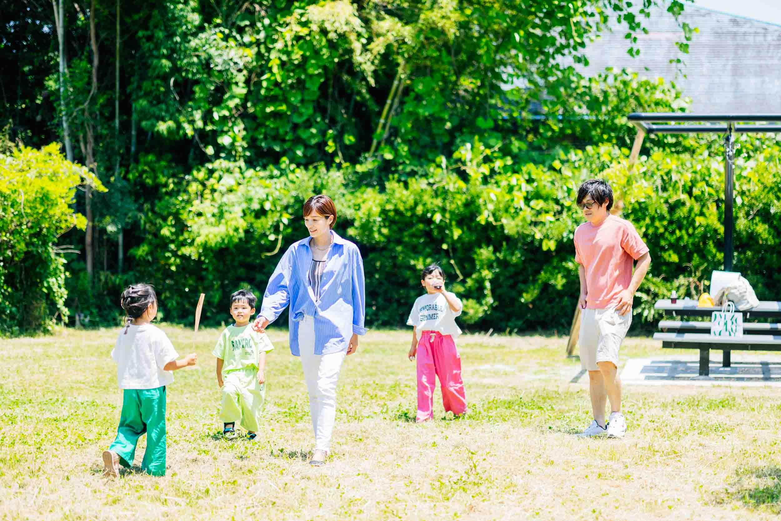 A family of five outdoors in a sunny park, walking on the grass with lush green trees in the background (mother, father, and three children).