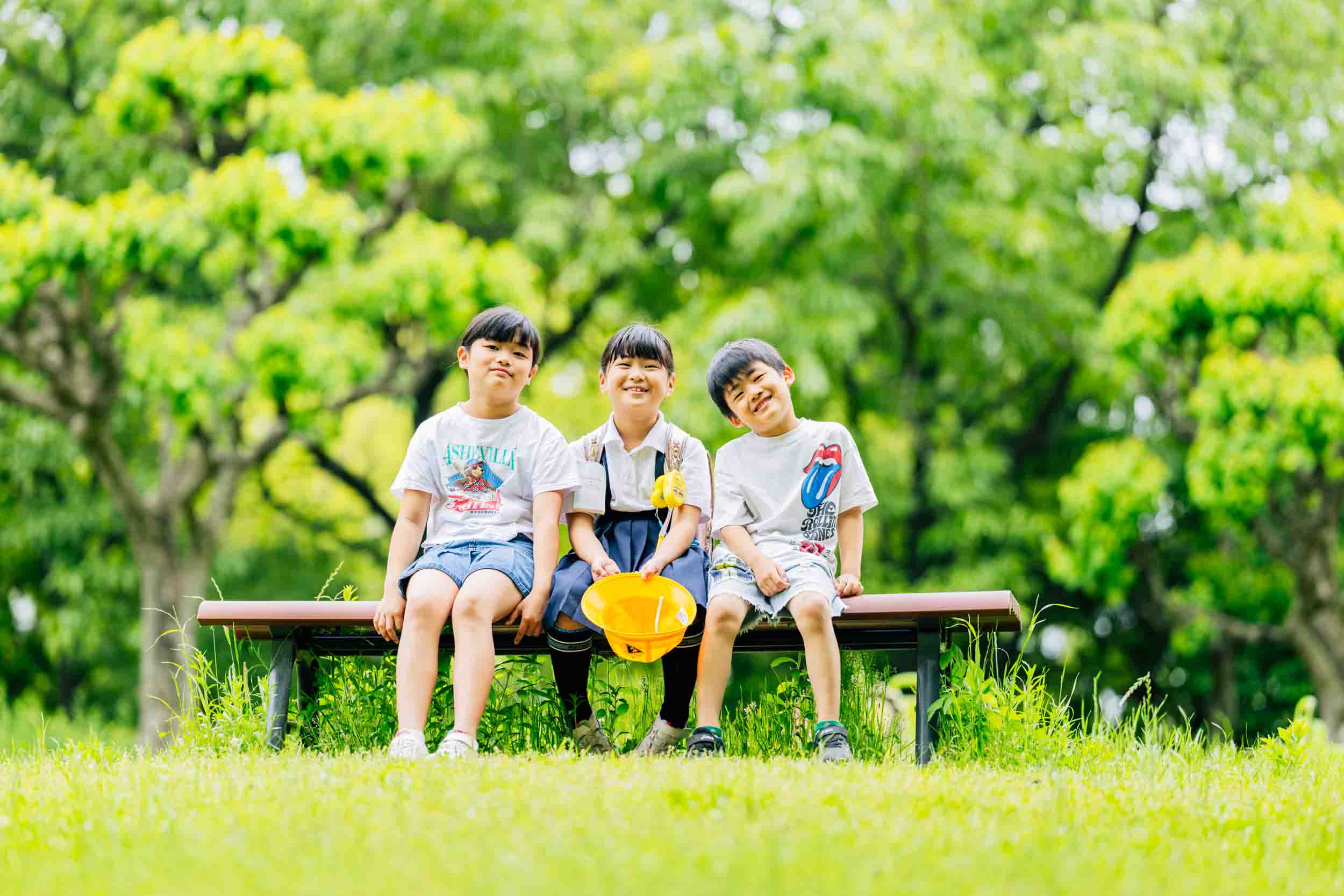 Three smiling children sit on a park bench with green trees behind them.