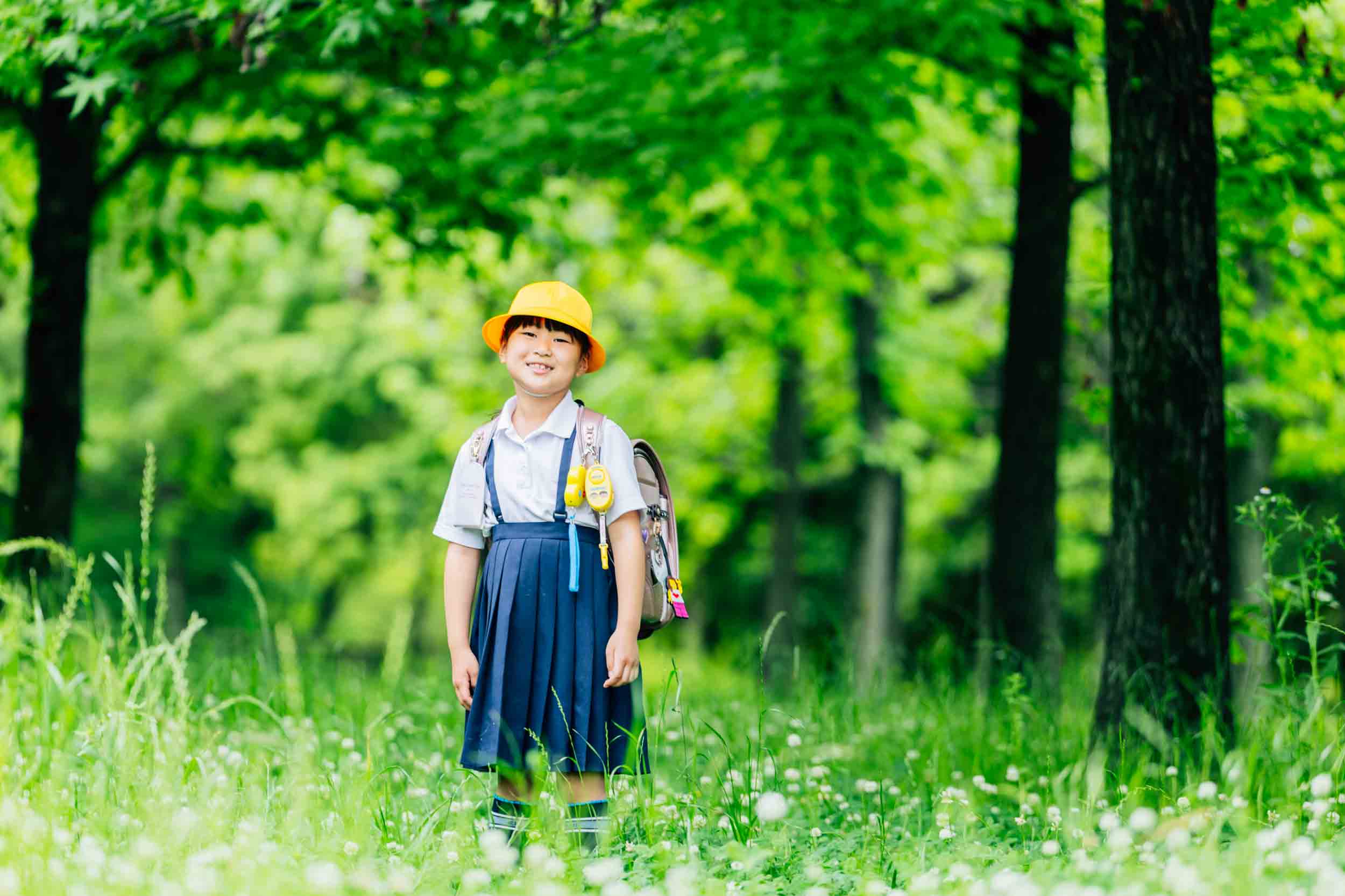 Smiling schoolgirl in a white shirt, blue uniform, and yellow hat with a backpack standing in a sunlit green forest clearing.