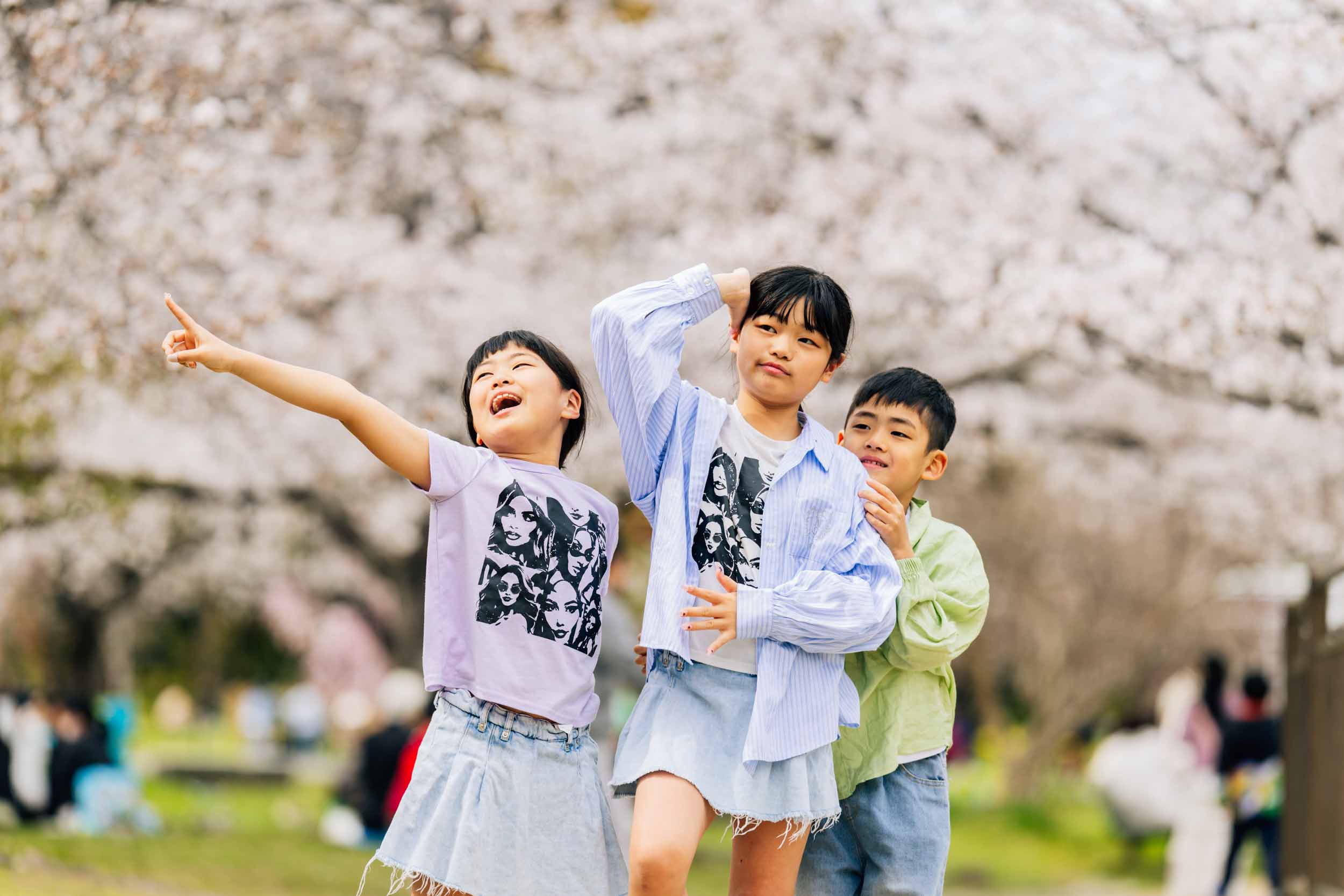 Three children playfully posing and dancing under blooming cherry blossoms in a park, smiling at the camera/around.