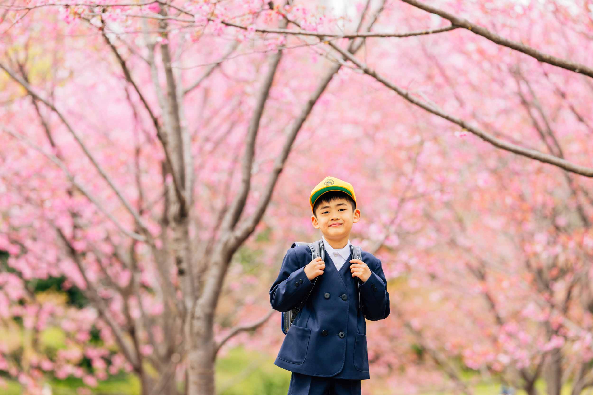 Boy in a navy school uniform with a yellow cap and backpack stands among pink cherry blossoms, smiling at the camera as blossoms surround him.