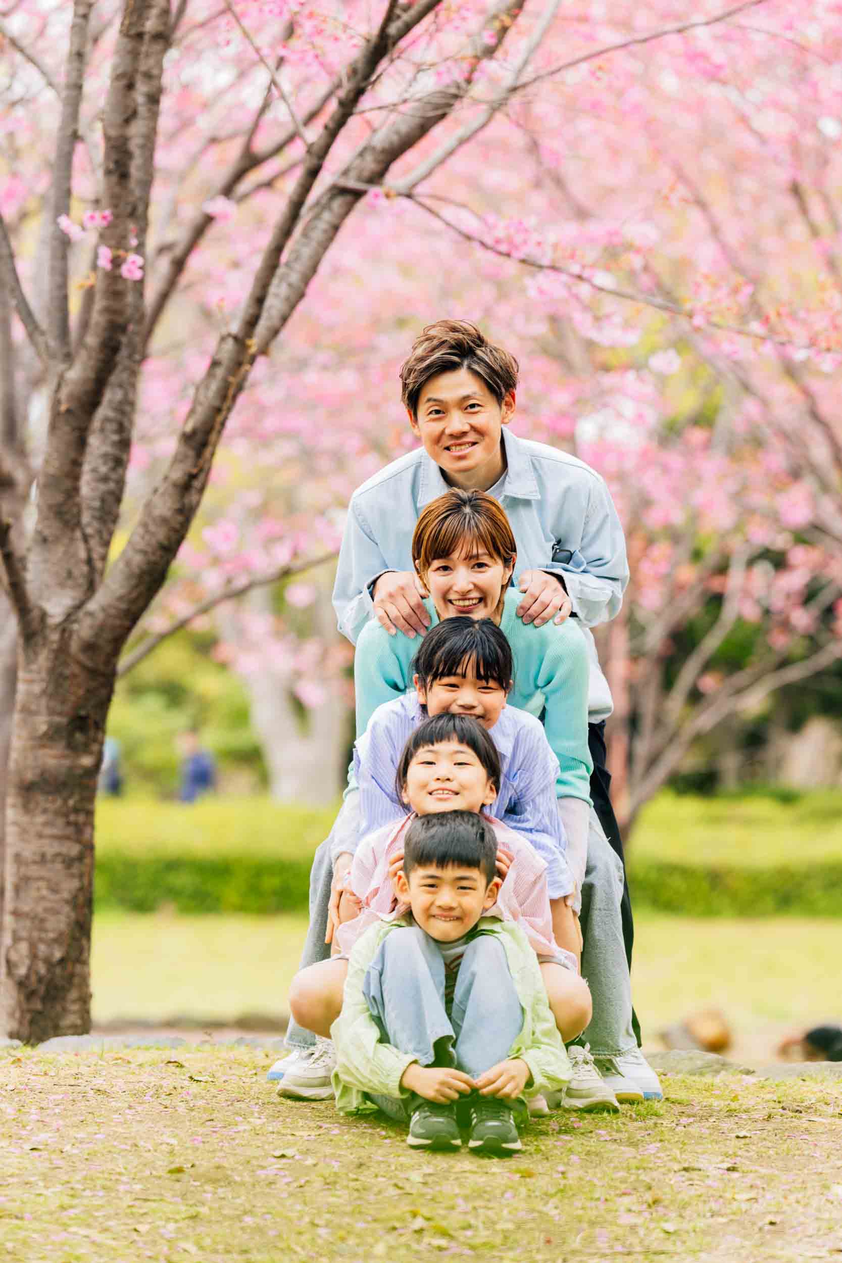 A family of five stacked in a human pyramid under pink cherry blossoms, smiling at the camera in a park.