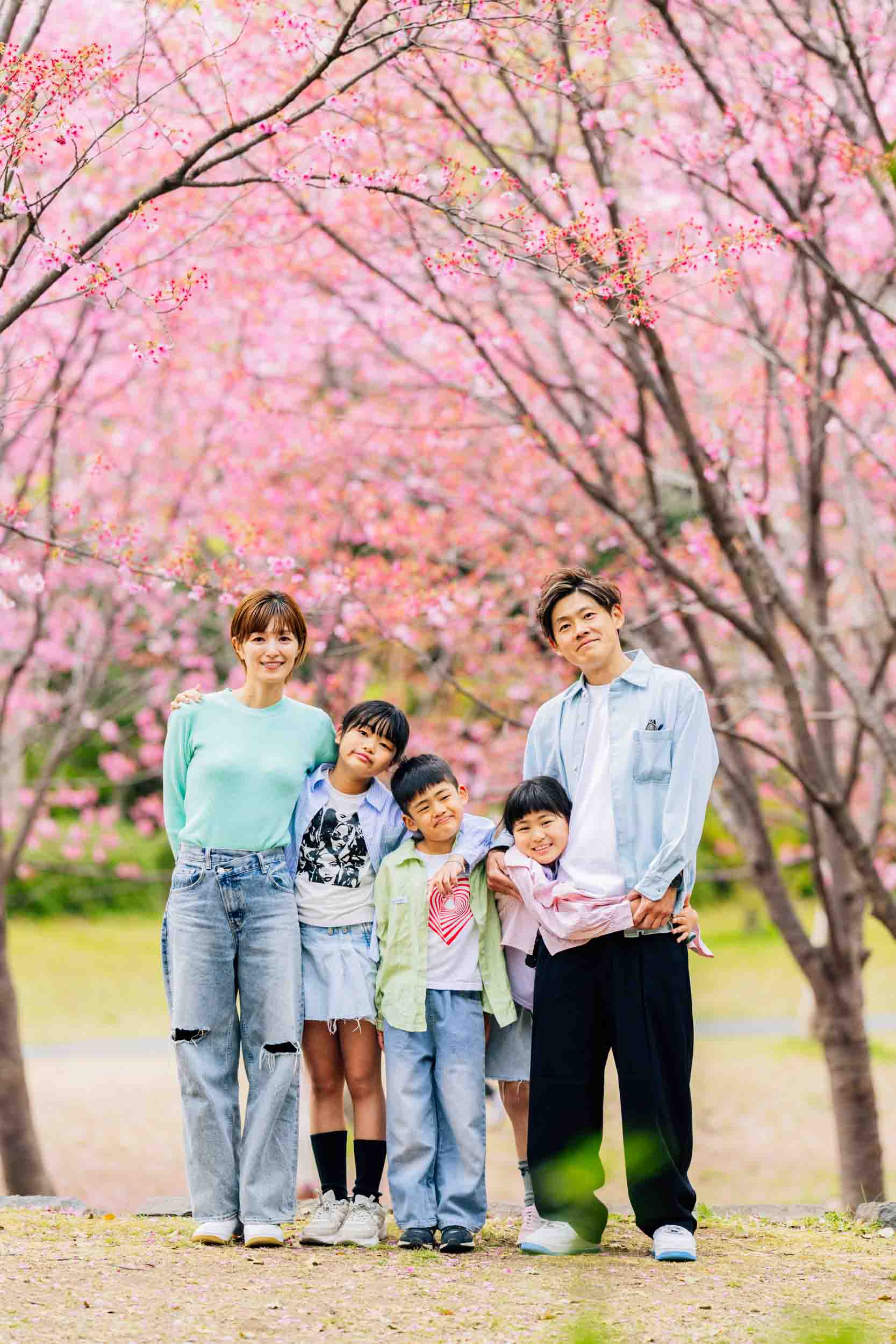 A family of five posing together under pink cherry blossom trees in a park, smiling at the camera.