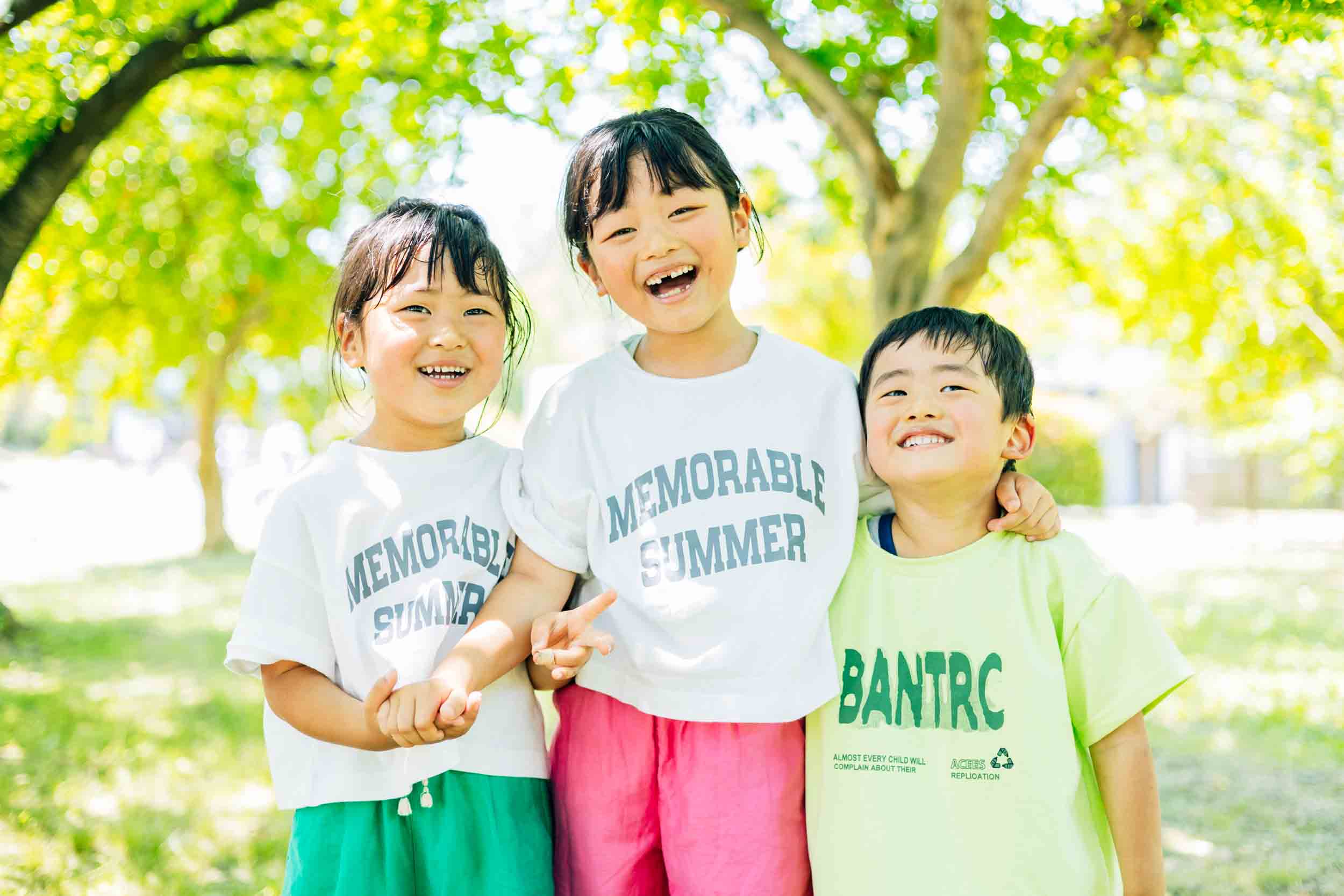 Three smiling children stand together outdoors in a sunny park, wearing matching white t‑shirts that read 'Memorable Summer' (left and center) and a lime shirt (right).