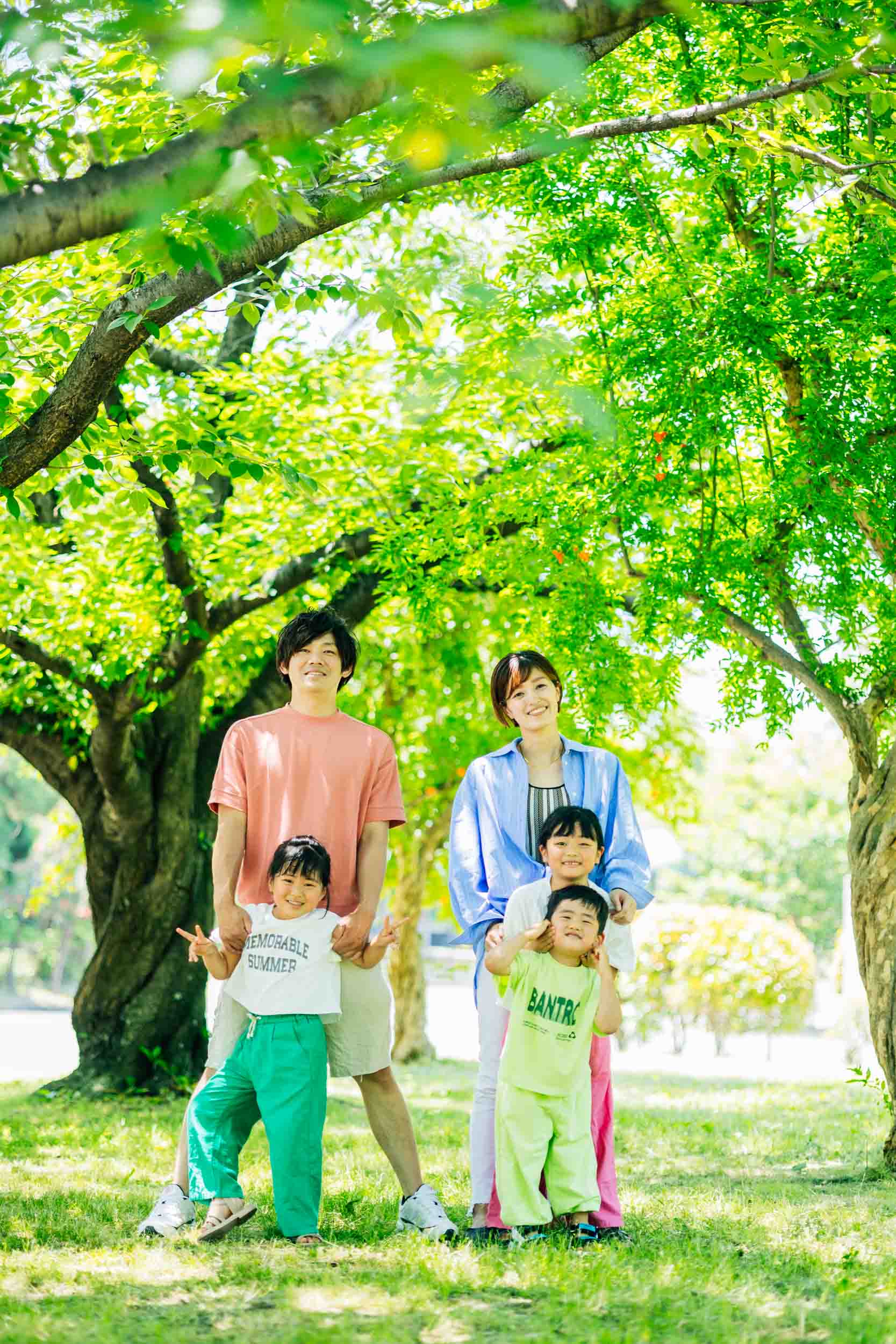 Family of four posing in a sunlit park under leafy trees with smiling parents and two kids nearby, casual clothing.