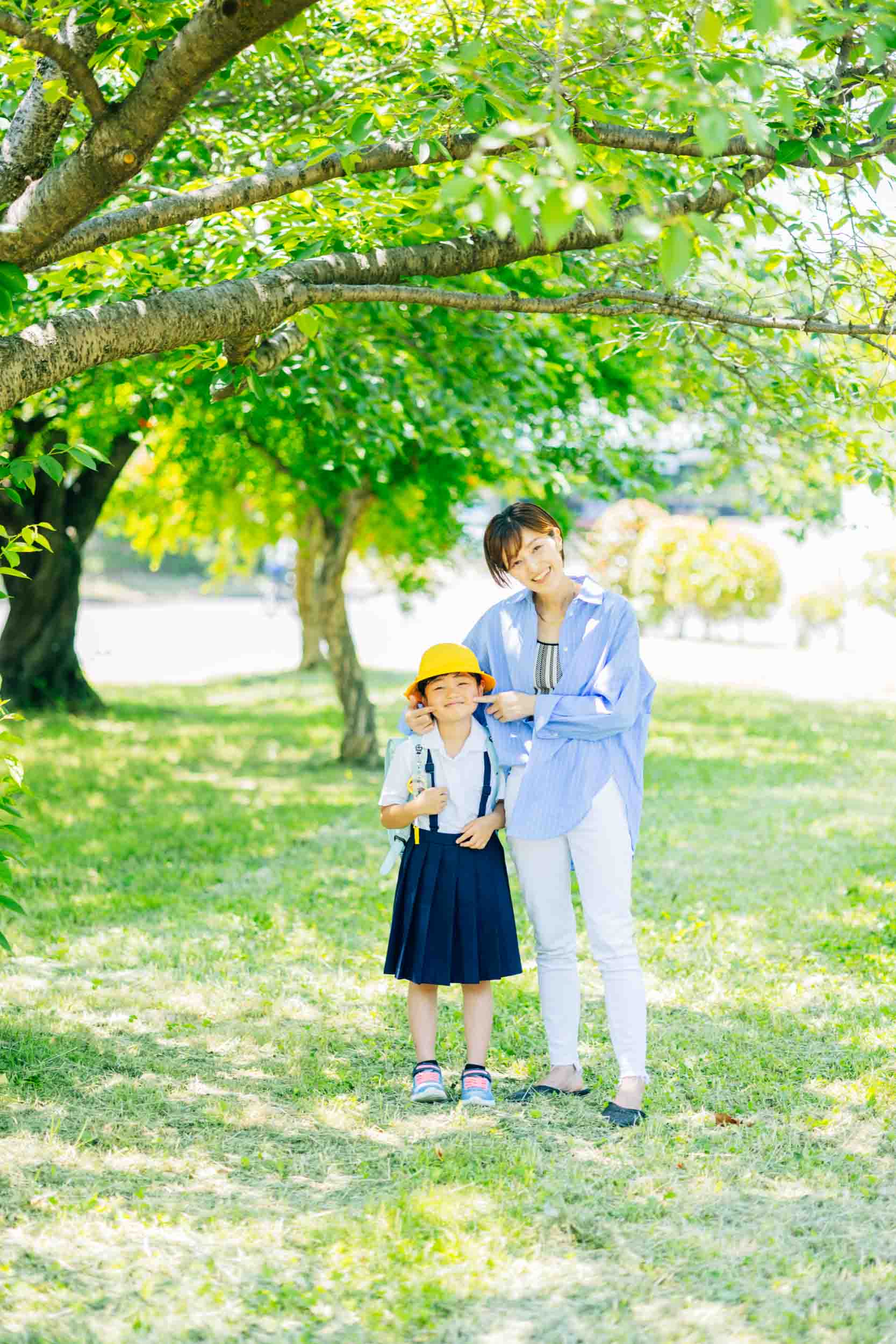 Mother adjusting her child’s yellow hat in a sunlit park with green trees and grass behind them, both smiling.