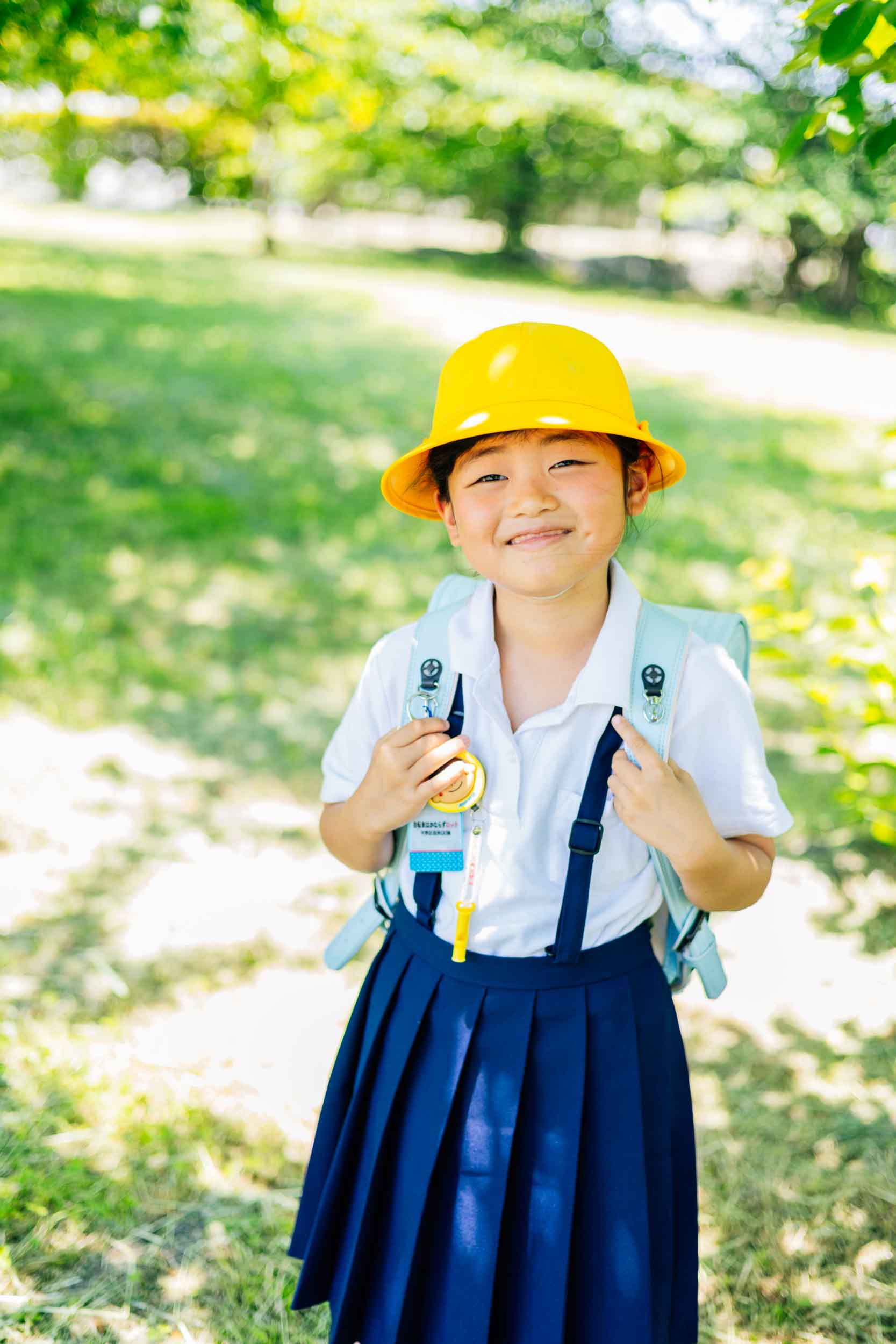 Smiling young girl in a white shirt and navy pleated skirt wearing a yellow hat and backpack outdoors in a sunny park.