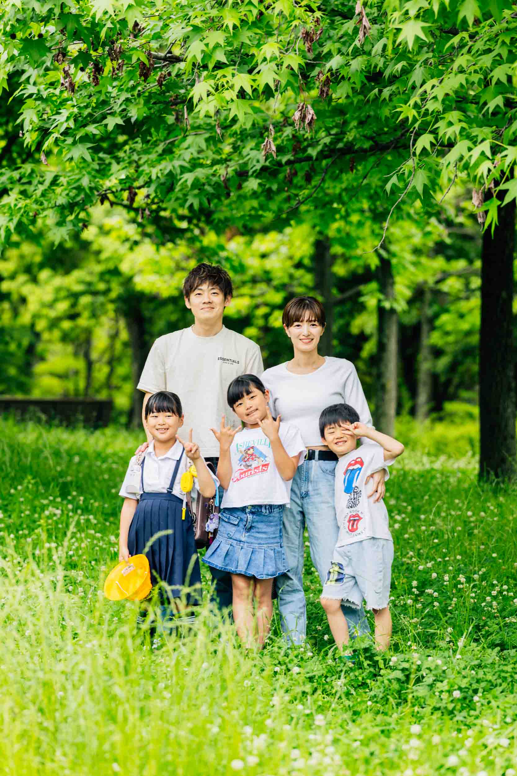 Family of four posing together in a lush, sunny park under leafy trees, smiling at the camera.
