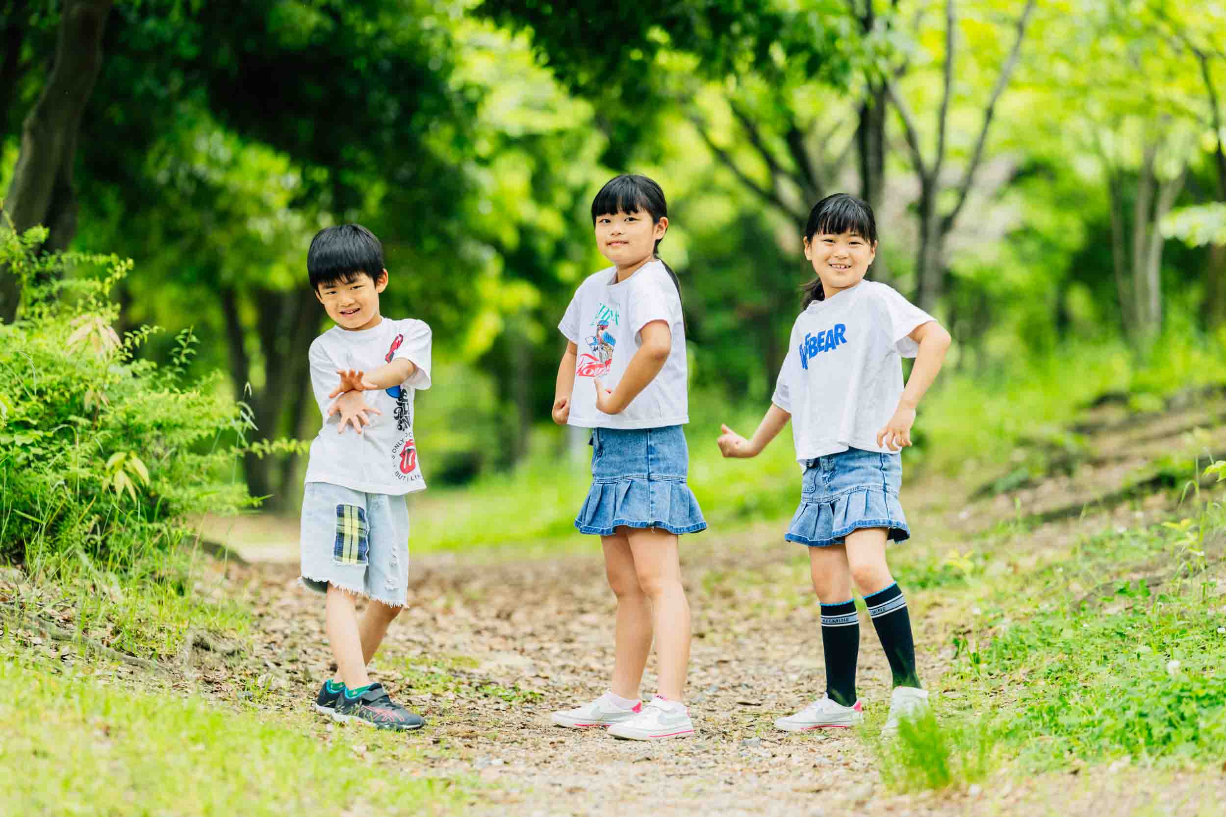 Three children in white T-shirts and denim outfits posing on a dirt path in a green park setting.