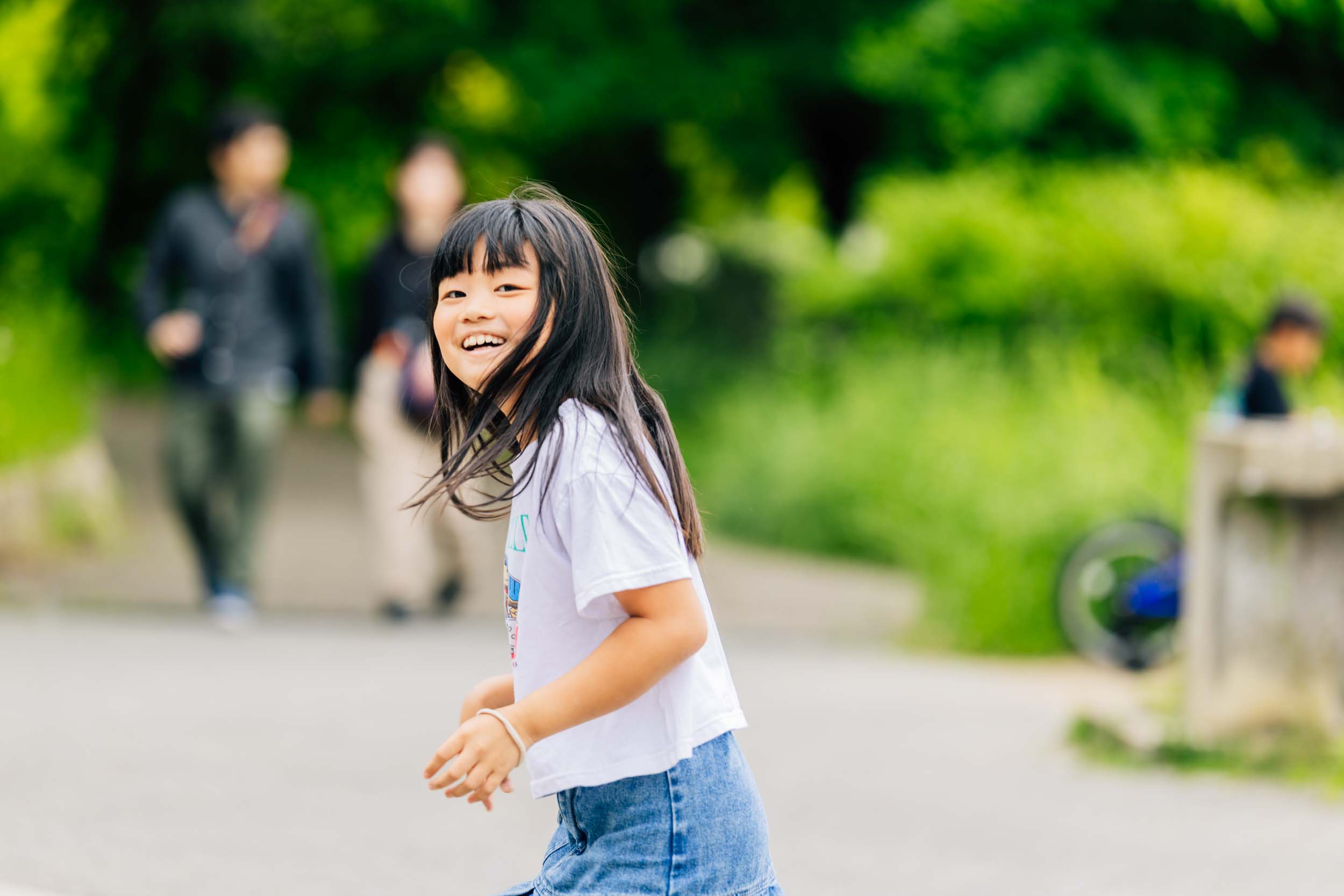 Smiling girl with long black hair runs toward the camera along a park path, wearing a white shirt and denim shorts/casual outfit.