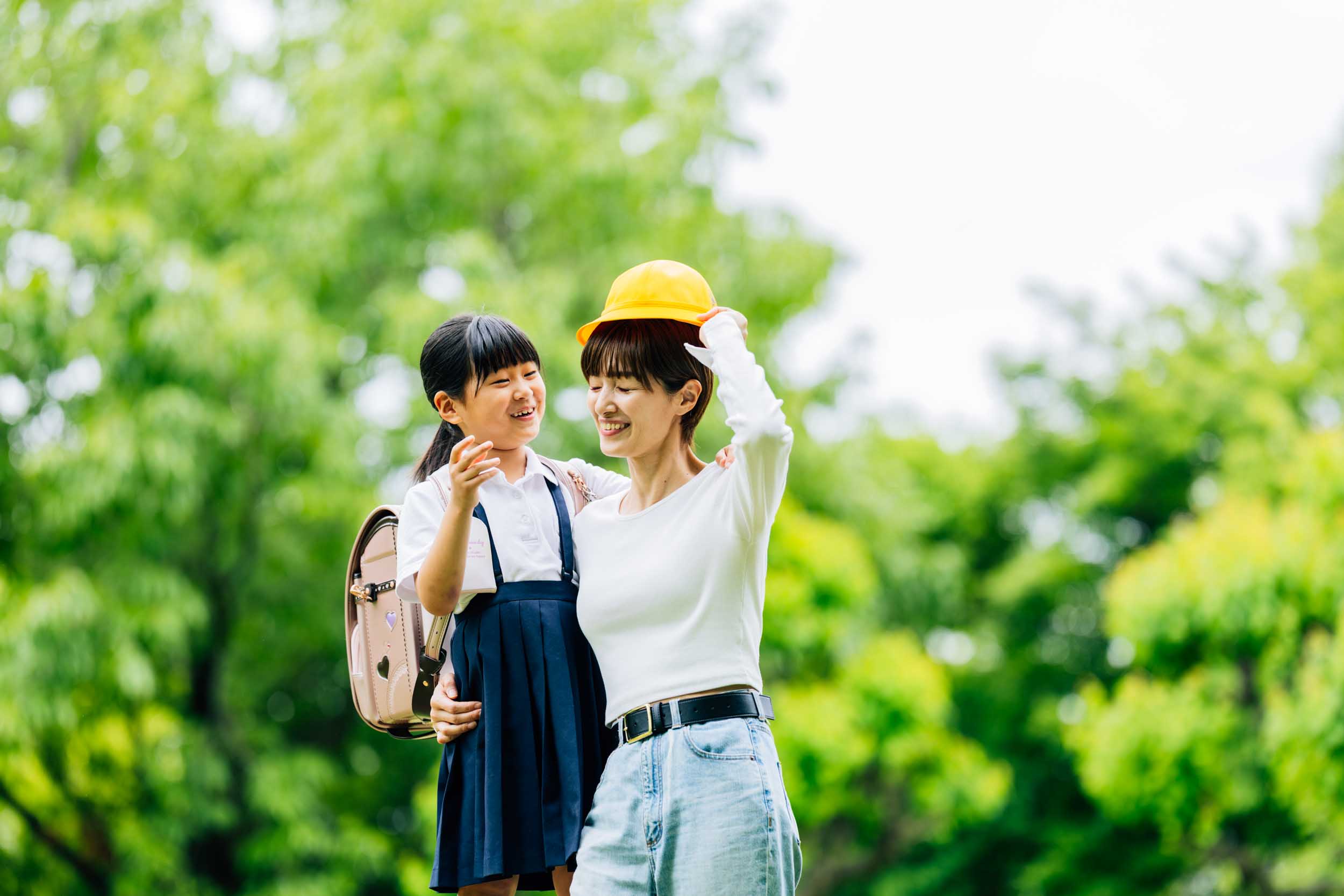 Mother and daughter share a joyful moment in a sunny green park; the girl wears a school uniform and backpack while the mom adjusts her orange hat.